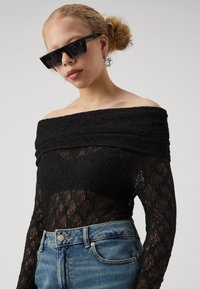 Top in pizzo nero senza spalle con motivo testurizzato, abbinato a jeans in denim a vita alta. Gli accessori includono occhiali da sole scuri e angolari.
