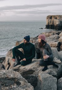 Zwei Personen sitzen auf felsigem Gelände in der Nähe des Wassers und tragen eng anliegende Outdoor-Bekleidung: ein dunkelgrünes Oberteil und eine Hose, ein graues Outfit und Strickmützen.