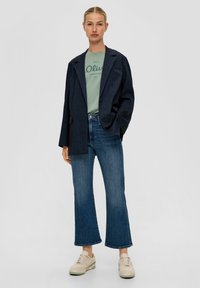 Marineblauwe gestreepte blazer over een lichtgroene graphic t-shirt, gecombineerd met uitlopende blauwe jeans en beige sneakers. Eenvoudig, casual outfitontwerp.