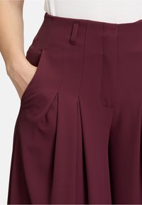 Pantaloni plissettati borgogna con tasche laterali e cuciture visibili lungo la vita. Realizzati in un tessuto liscio e leggero.
