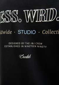 Black cotton t-shirt featuring white and light blue printed text, with various fonts stating "ESSENTIALS," "STUDIO," and "Worldwide Collection."