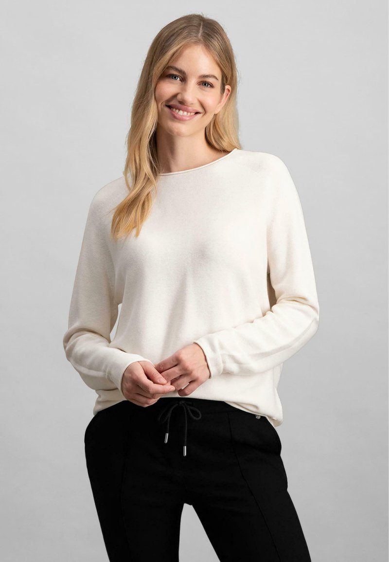 Helles beiges Sweatshirt mit langen Ärmeln und rundem Ausschnitt, kombiniert mit schwarzen Hosen mit Zugband und silbernen Akzenten.