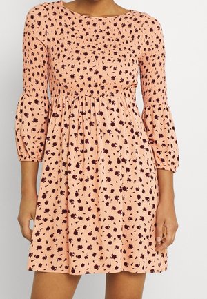 Frau trägt ein pfirsichrosa Kleid mit Blumenmuster, gerafftem Oberteil und dreiviertel Puffärmeln, steht vor einem einfarbigen weißen Hintergrund.