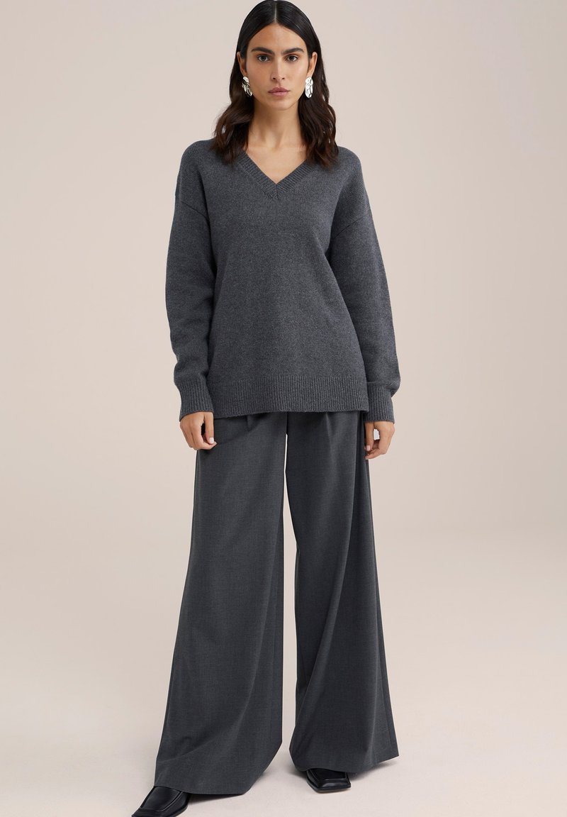 Grijze gebreide trui met een v-hals en een losse pasvorm, gecombineerd met brede grijze broeken. Soepele textuur en contrasterende ribbels bij de mouwen.