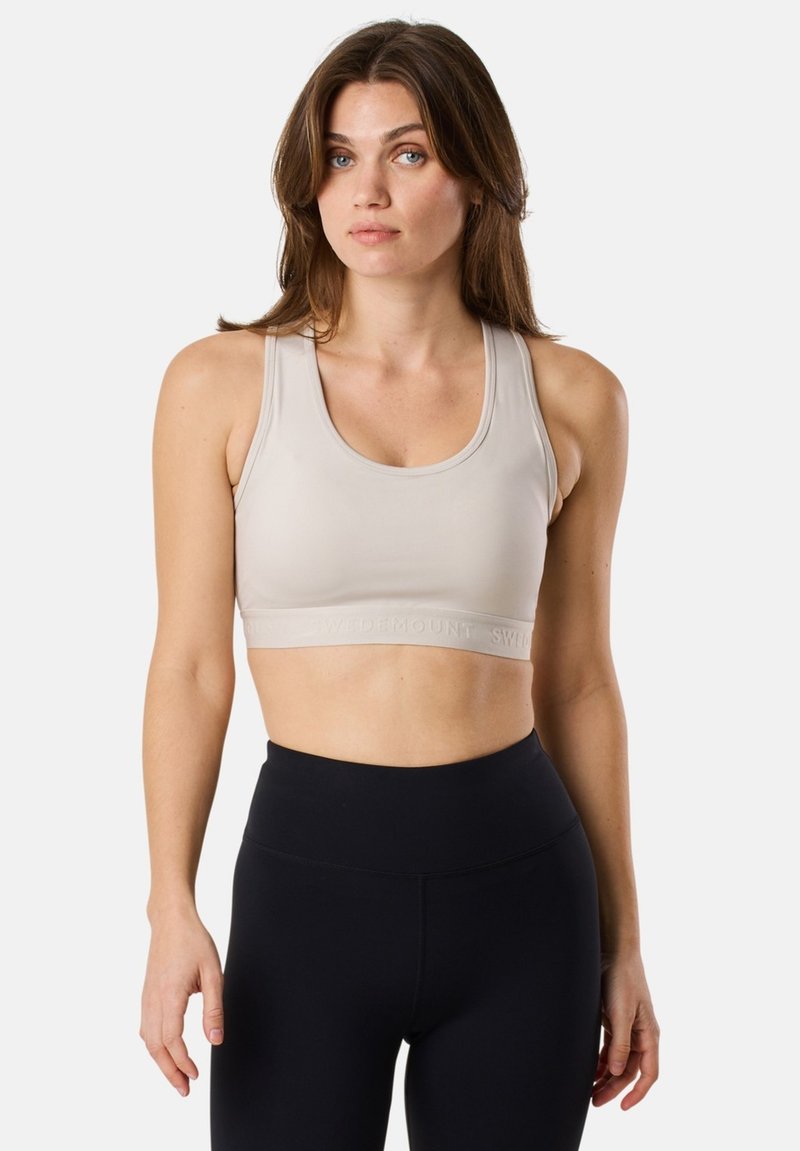 Soutien-gorge de sport beige avec un encolure ronde et des bretelles larges, présentant une texture solide et une bande logo en relief, associé à un legging noir taille haute.