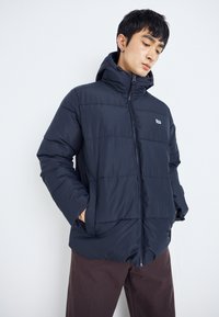 Veste matelassée navy avec une fermeture éclair à l'avant, une capuche et un motif matelassé horizontal. Elle dispose de poches latérales et d'un logo sur la poitrine. Texture lisse.