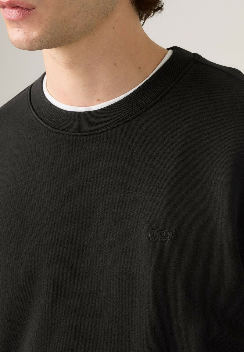 Jeune personne portant un sweat-shirt noir Levi’s sur un maillot blanc, coupé au niveau du menton et des épaules.