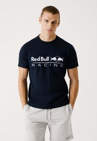Tamsiai mėlynas medvilninis marškinėliai su balta "Red Bull Racing" užrašu ir logotipu. Trumpomis rankovėmis, apvaliu kaklu, vilkima su šviesiai pilkais šortais.