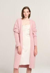 Langer, offener Cardigan aus weichem, rosa Strick mit übergroßen Ärmeln und geripptem Saum. Getragen über einem cremefarbenen Satinkleid.