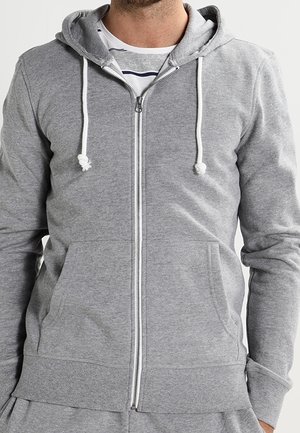 Hombre vistiendo una sudadera con cremallera gris con cordones blancos sobre una camiseta a rayas blanca y gris.