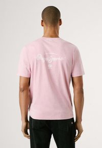 Camiseta de manga corta rosa claro con cuello redondo, con un diseño de logo blanco en la parte posterior. Fabricada con suave tela de algodón.