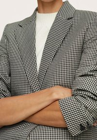 Czarnobiały żakiet w wzór houndstooth z wciętymi klapami, teksturowanym materiałem i zapinanymi mankietami. Nałożony na biały sweter z prążkowanej dzianiny.
