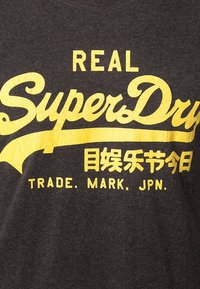 Camiseta gris con un atrevido texto amarillo que dice "REAL SuperDry" y un logotipo adicional en una fuente más pequeña.