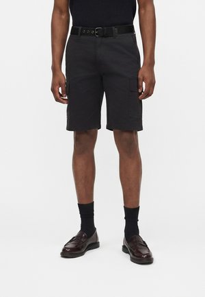 Άτομο που φοράει μαύρα cargo σορτς με πλαϊνές τσέπες, μαύρη ζώνη, μαύρες κάλτσες και καφέ δερμάτινα loafers, στέκεται μπροστά σε απλό φόντο.