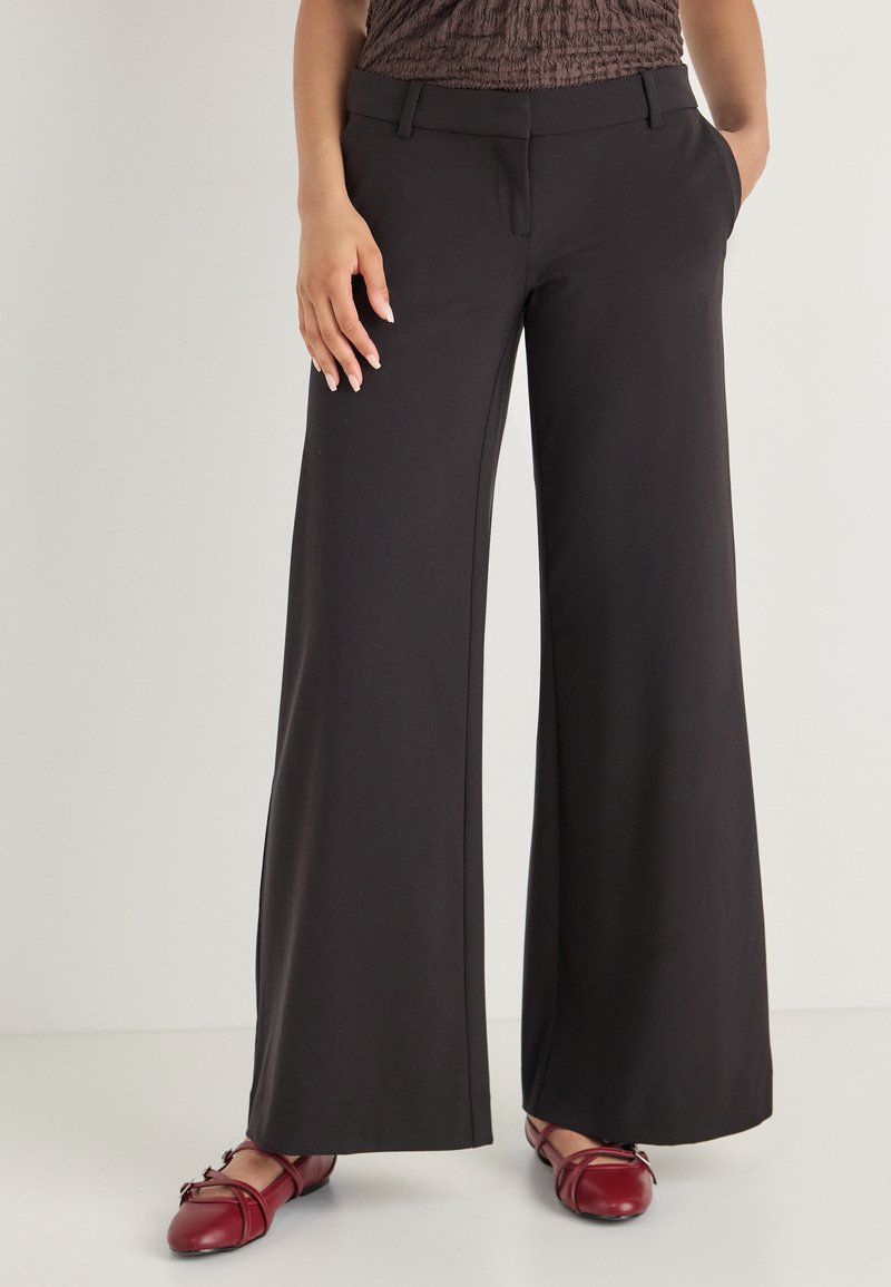 Pantalons noirs à jambes larges en tissu lisse ; dotés de poches latérales et d'une taille ajustée. Associés à des ballerines bordeaux.