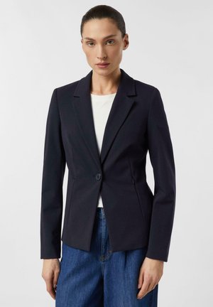 Kobieta ubrana w dopasowaną czarną marynarkę na białą bluzkę i niebieskie dżinsy, stojąca na tle jednolitego jasnego tła.