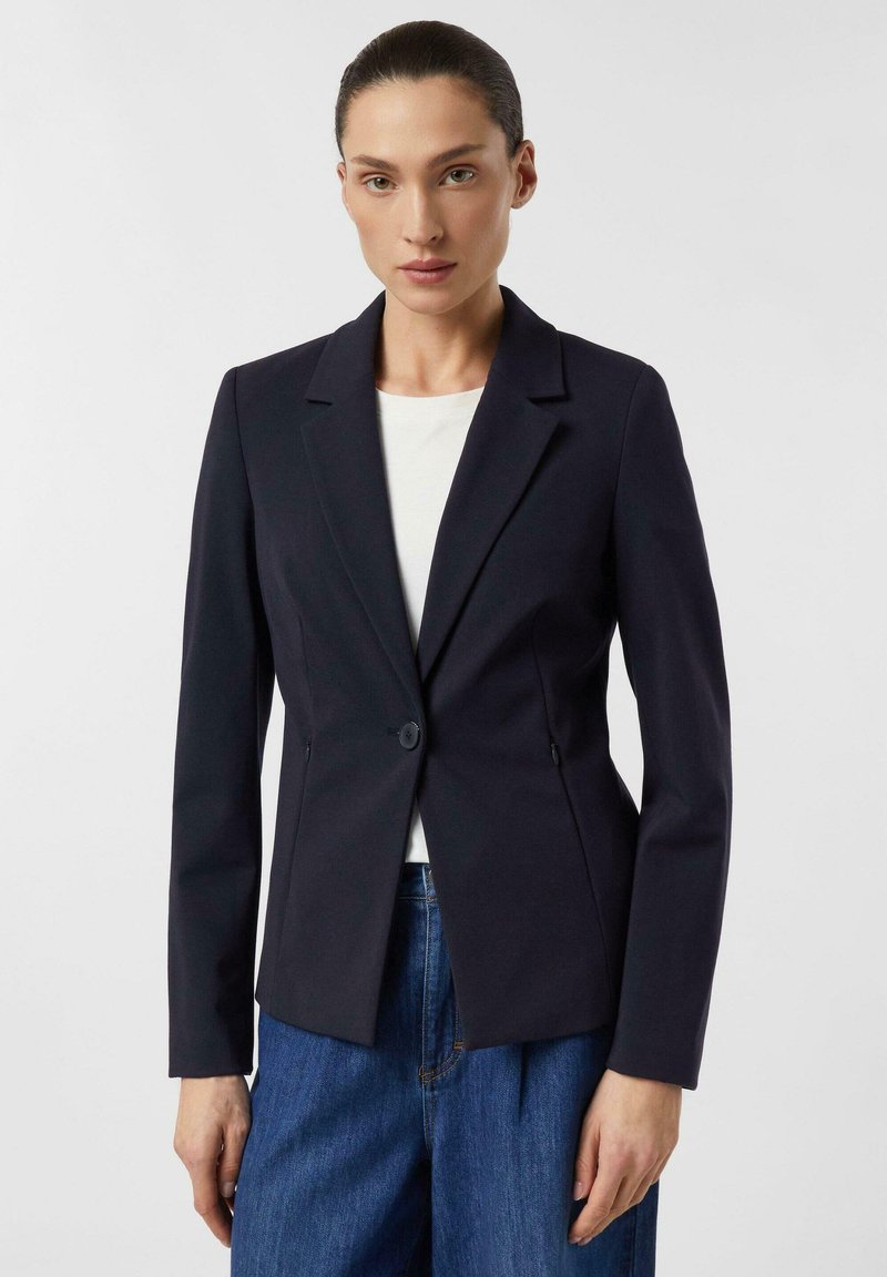 Frau trägt ein eng anliegendes schwarzes Sakko über einem weißen Oberteil und blauen Jeans, steht vor einem schlichten hellen Hintergrund.