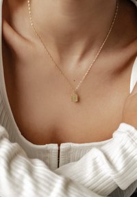 Collier en pendentif en or sur une chaîne fine, doté d'un charme rectangulaire avec une surface texturée, présenté sur un haut de couleur claire.