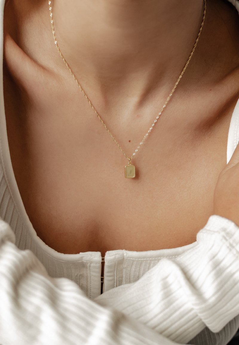 Collier en pendentif en or sur une chaîne fine, doté d'un charme rectangulaire avec une surface texturée, présenté sur un haut de couleur claire.