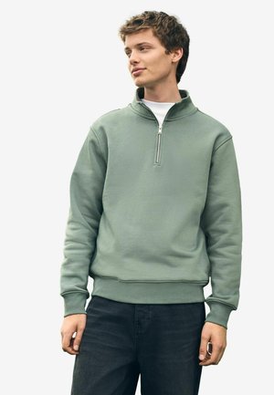 Green, zip-up sweatshirt with a textured finish, ribbed cuffs, and hem. Worn over a white shirt, paired with dark denim pants.