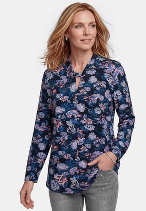 Frau mit blonden Haaren, die eine langärmelige, dunkelblaue, florale Bluse und graue Jeans mit Strasssteindetails trägt und mit einer Hand in der Tasche posiert.