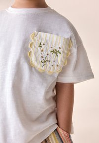 Fehér póló hullámos sárga csíkos zsebbel, virágos hímzéssel díszítve. Puha anyag; rövid ujjú; laza szabás.