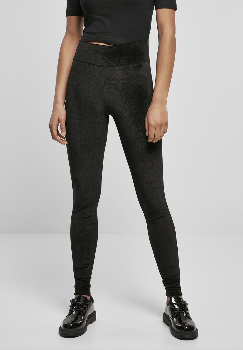 Kvinna som bär svarta åtsittande sammetsleggings, en kort svart topp och glänsande svarta snörskor, stående mot en grå bakgrund.