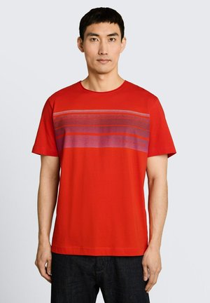 Mężczyzna w czerwonej koszulce z krótkim rękawem z poziomymi fioletowymi paskami na klatce piersiowej, w zestawieniu z ciemnymi spodniami.