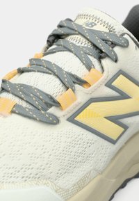 Gros plan d'une basket blanche avec des lacets gris, des passants jaunes, un logo "N" jaune et gris, et une semelle texturée.