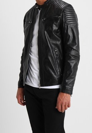 Mężczyzna ubrany w czarną pikowaną skórzaną kurtkę na białej koszulce i czarnych spodniach, stojący na jednolitym tle.