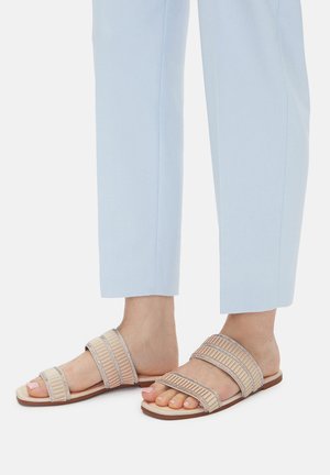 Beżowe sandały na płaskiej podeszwie z trzema teksturowanymi paskami ozdobionymi metalicznymi akcentami, noszone z jasnoniebieskimi, krótkimi spodniami, odsłaniające palce.