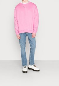 Ljust rosa sweatshirt med rund halsringning, kombinerad med ljusblå jeans och vita skor med svarta sulor och snörningar.