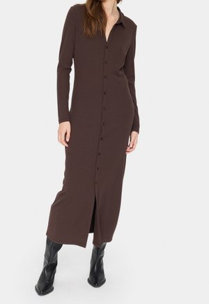 Femme portant une longue robe boutonnée côtelée marron foncé à manches longues et des bottes noires pointues sur fond blanc.
