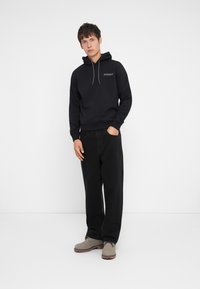 Black hoodie with a drawstring, featuring a subtle logo on the chest, paired with loose black pants and light gray ankle boots.