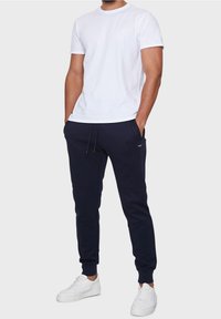 T-shirt en coton blanc associé à un pantalon de survêtement marine avec cordon de serrage. Le modèle porte des baskets blanches. Texture lisse, design décontracté.