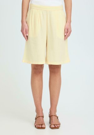 Kvinne iført knelangt, lyst gule, løstsittende shorts og brune sandaler med hæl, stående foran en ensfarget bakgrunn.