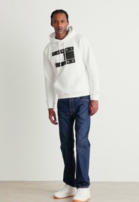Sudadera blanca con el logo negro "TOMMY JEANS", combinada con jeans de mezclilla azules y zapatillas blancas con suela de color marrón. Textura de tela suave.