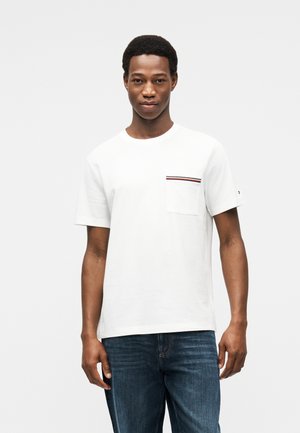 Jeune homme portant un t-shirt blanc à manches courtes avec une petite poche poitrine et un jean bleu, debout devant un fond clair uni.