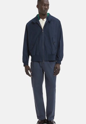 Uomo che indossa una giacca blu navy con zip, camicia bianca con colletto e rifiniture verdi, pantaloni blu navy abbinati e scarpe scure, in piedi davanti a uno sfondo semplice.