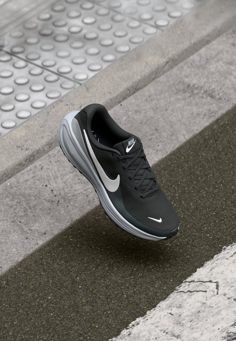 Scarpa da corsa nera Nike con una parte superiore testurizzata, suola grigia e logo swoosh bianco. La scarpa è posizionata sopra una superficie in cemento.