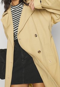 Helle beigefarbene Trenchcoat mit großen Knöpfen, ausgestattet mit einem Reverskragen und Seitentaschen, über einem schwarzen Denimrock und einem gestreiften Oberteil getragen.