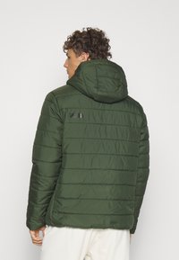 Giacca puffer verde con una texture trapuntata, cappuccio e un accento del logo nero sul retro. Caratterizzata da una vestibilità rilassata e un design lineare.