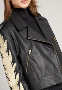 Vrouw draagt een zwarte leren jas met gouden ritsen en beige studs in vlammenmotief op de mouw, over een zwarte coltrui.