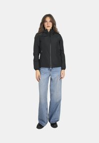 Giacca nera con zip e colletto alto, caratterizzata da maniche lunghe e un design aderente. Abbinata a jeans a gamba larga di un blu chiaro e scarpe nere.