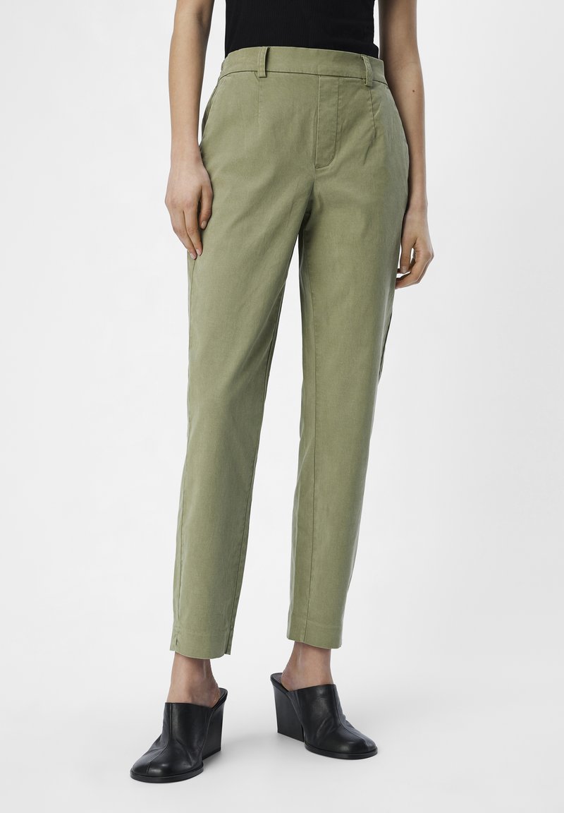 Pantalons fuselés vert kaki avec des plis à l'avant, des poches latérales et une texture lisse. Associés à des mules à talons noirs pour le contraste.