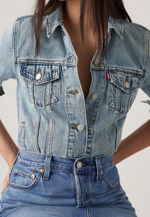 Woman wearing light blue denim jacket with silver buttons and blue high-waisted denim jeans, hands resting on hips.