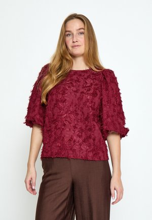 Žena s dugom plavom kosom, odjevena u teksturiranu bordo bluzu s nabranim rukavima i široke smeđe hlače, stoji ispred jednobojne pozadine.