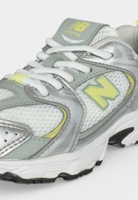 Γκρι, λευκό και κίτρινο παπούτσι New Balance για τρέξιμο με ύφασμα από δίχτυ, κορδόνια και υφασμένο πέλμα, εμφανιζόμενο από κοντινή γωνία.