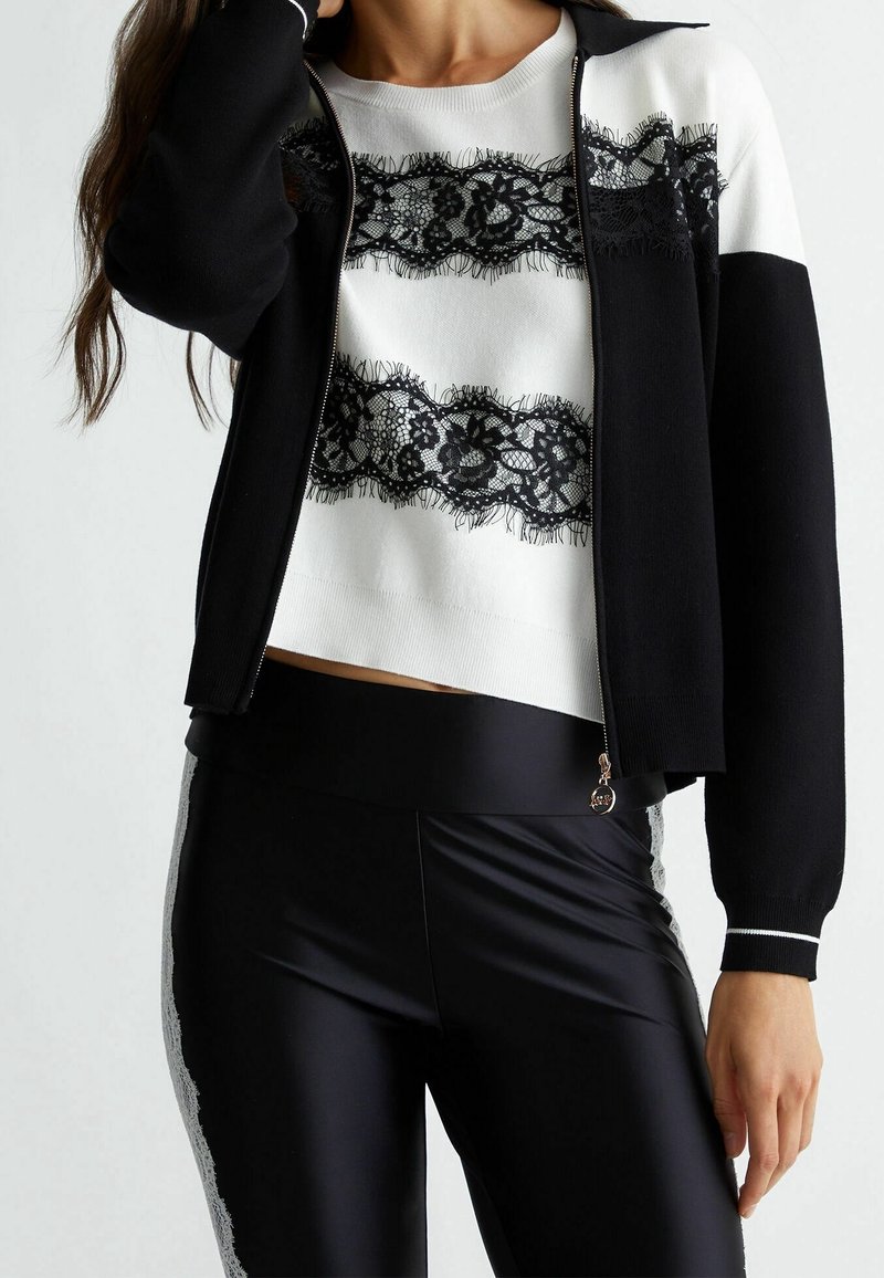Black zip-up jacket over a white sweater featuring black lace stripes, paired with form-fitting black pants with a lace side pattern.