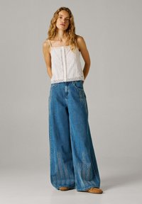 Mujer joven con cabello largo y ondulado, vestida con una blusa blanca sin mangas y pantalones vaqueros azules de pierna ancha bordados, posando frente a un fondo liso.